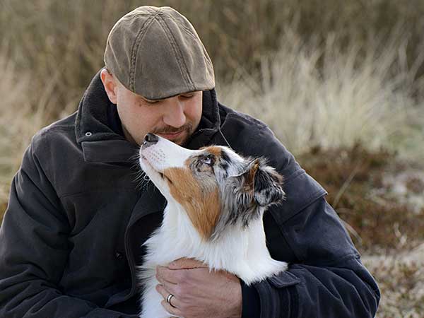 The Importance of Dog Behavior Training with Australian Shepherds - Photo: Man hugging Australian Shepherd outdoors.